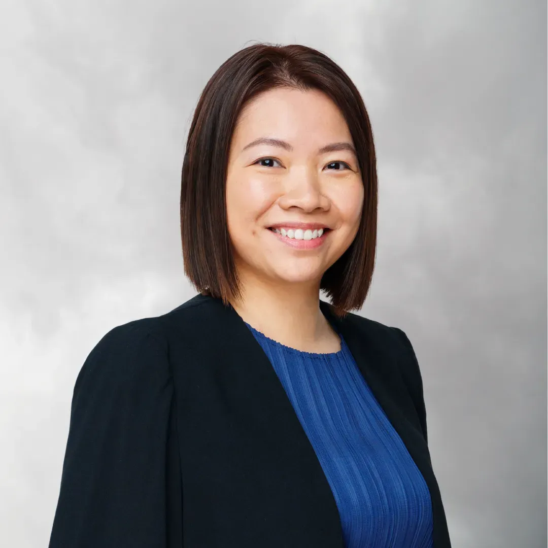 A smiling woman with shoulder-length dark brown hair, wearing a black blazer and a blue top, posed against a gray background.