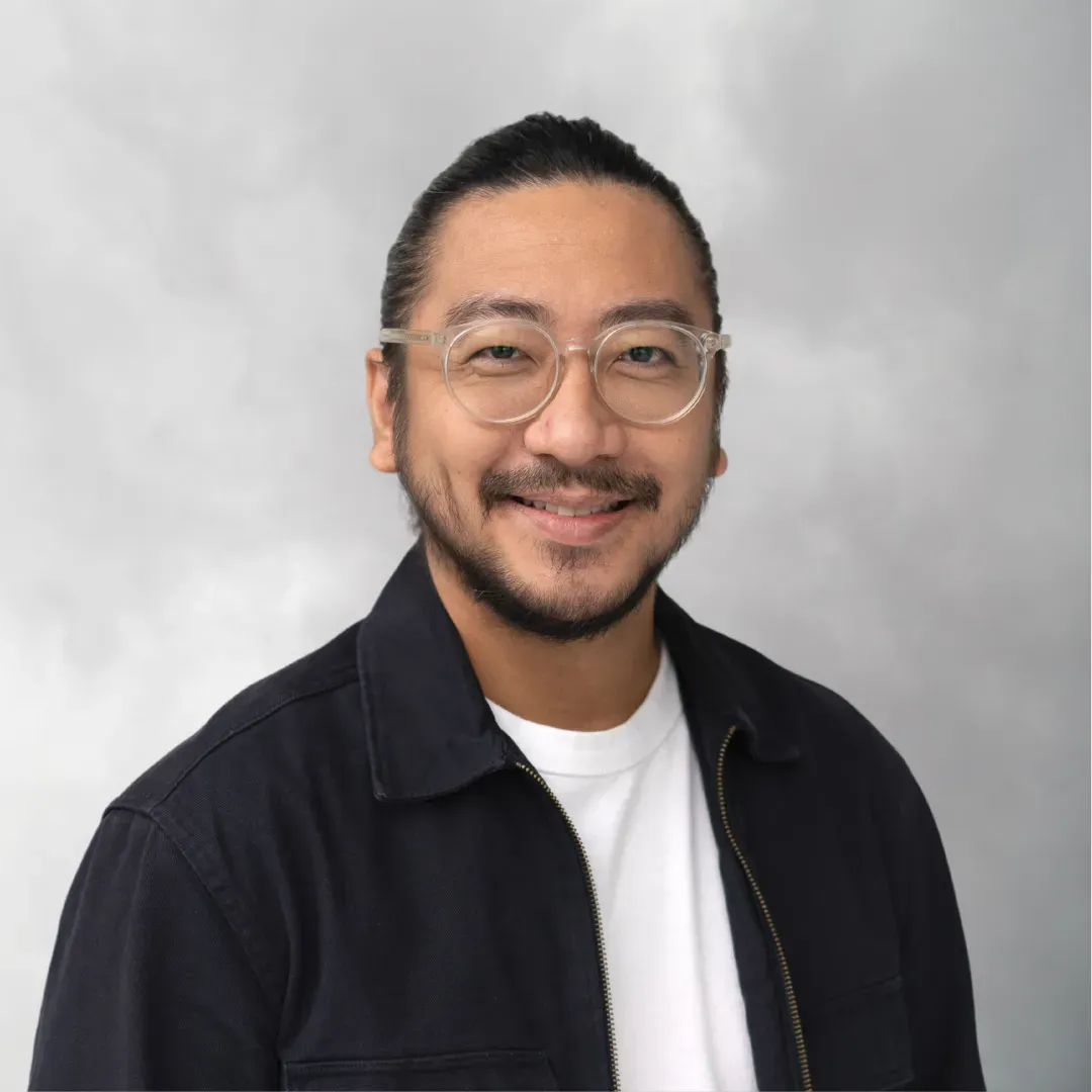 A man with glasses, a beard, and a bun hairstyle, smiling, wearing a black jacket and a white T-shirt, standing against a cloudy gray background.