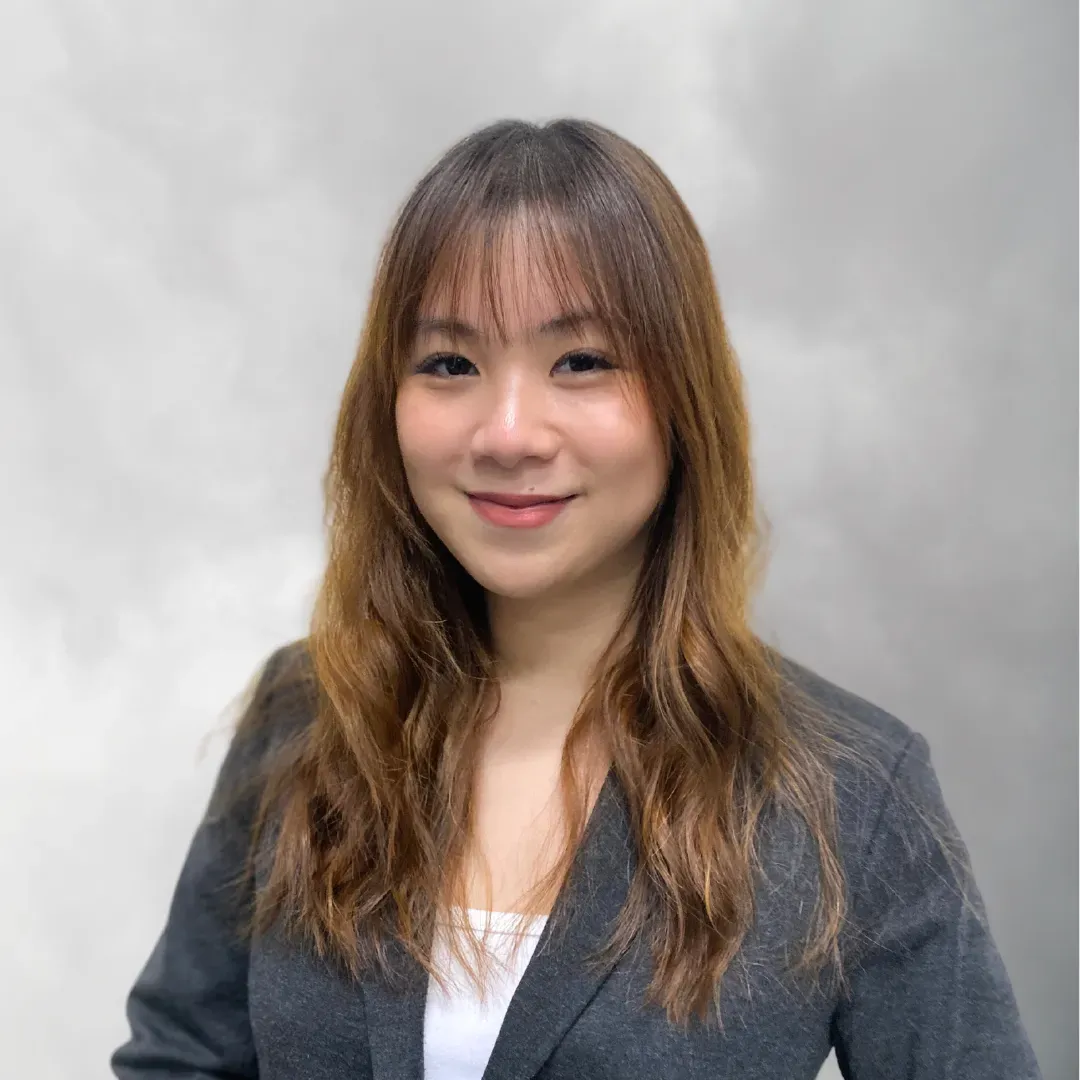 Portrait of a young woman with shoulder-length wavy hair, smiling, wearing a dark blazer over a white top, standing against a cloudy sky background.