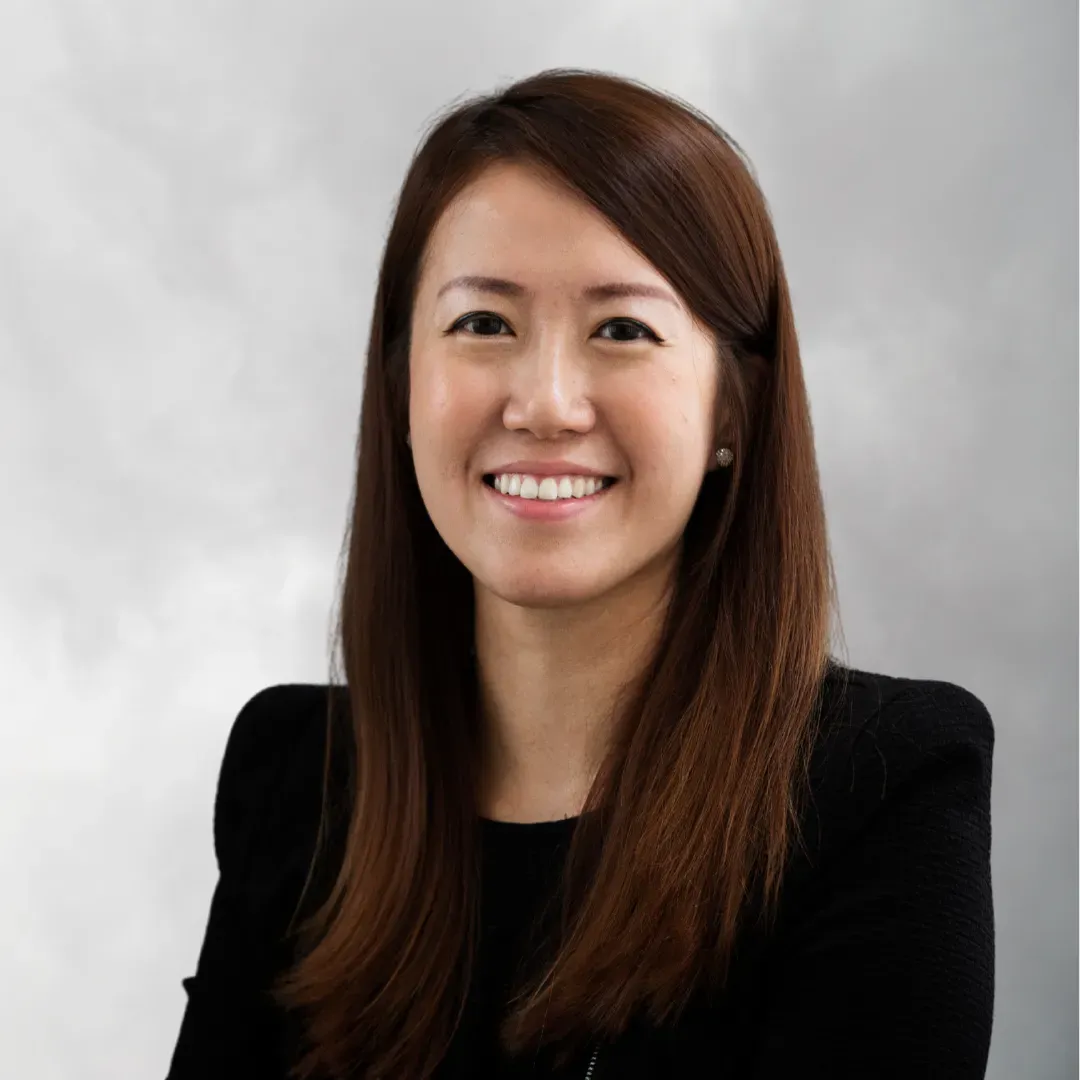 Portrait of a smiling woman with long brown hair, wearing a black top, against a gray background.
