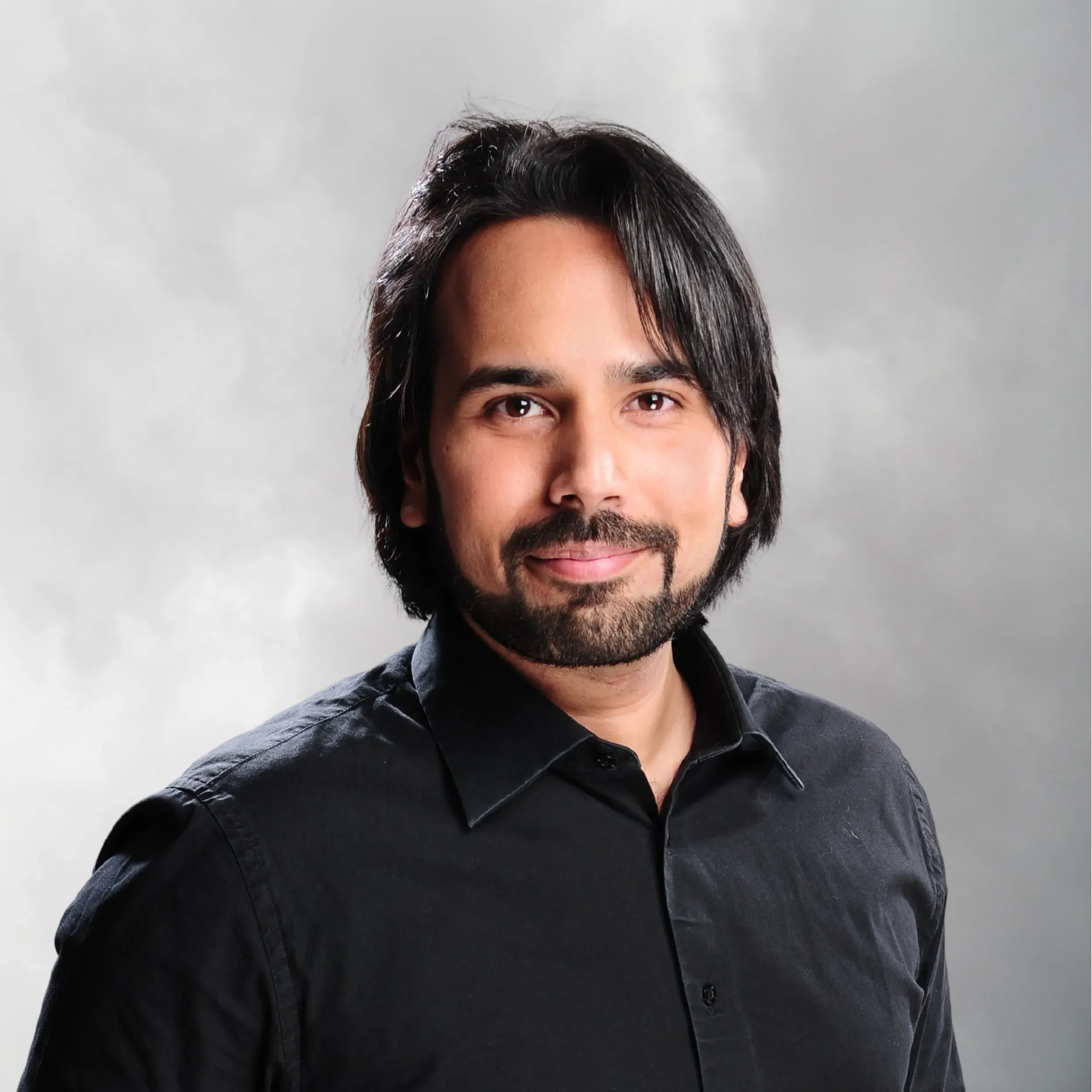 Headshot of a man with dark hair, a beard, wearing a black collared shirt, smiling against a neutral gray background.
