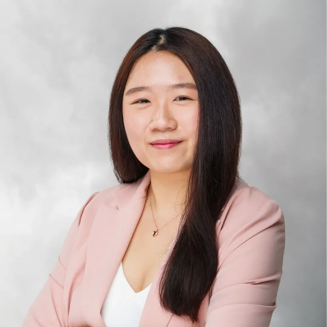 A woman with long dark hair, wearing a pink blazer and a white top, smiling slightly against a neutral gray background.