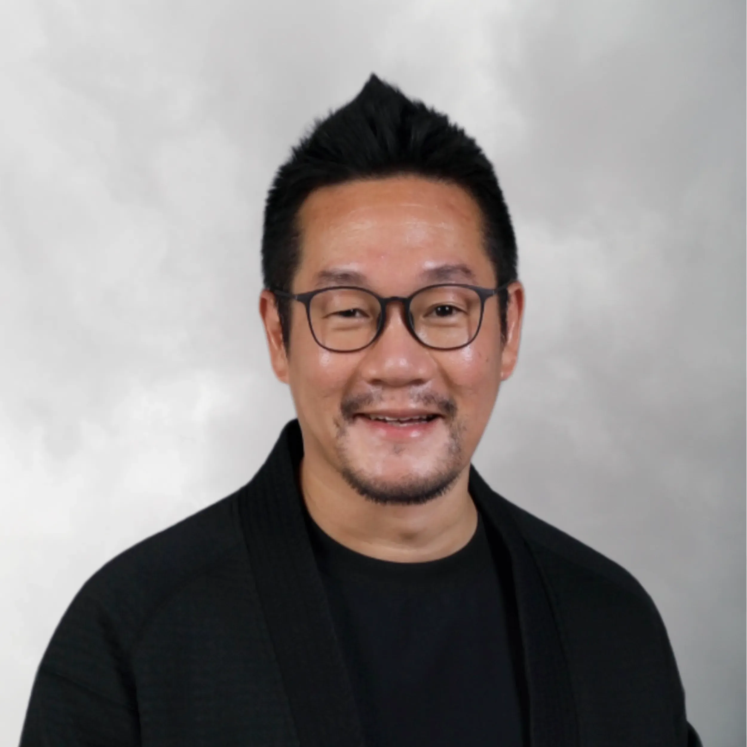 A smiling man with glasses, facial hair, wearing a black jacket against a plain light gray background.