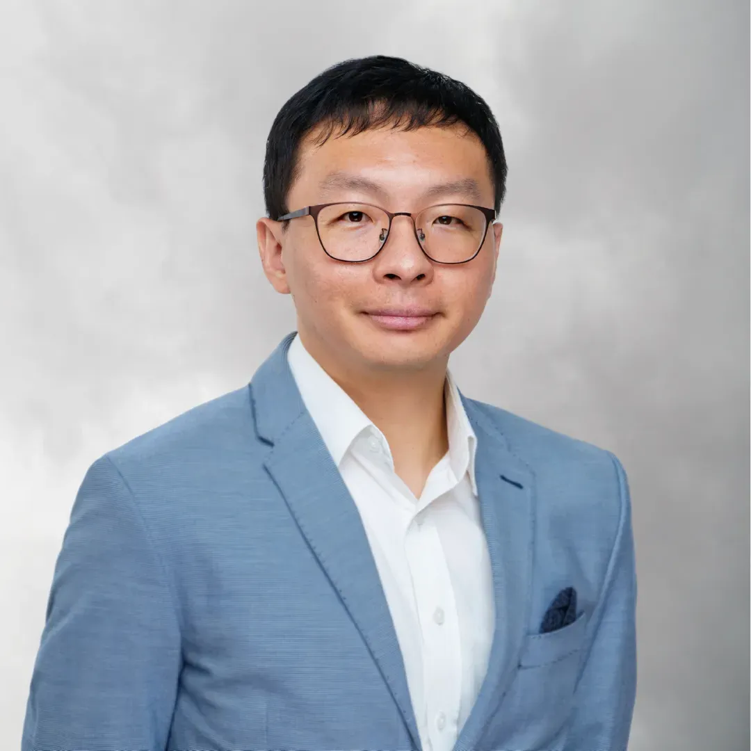 Portrait of a young man with short black hair and glasses, wearing a light blue blazer and white shirt, standing against a gray background.
