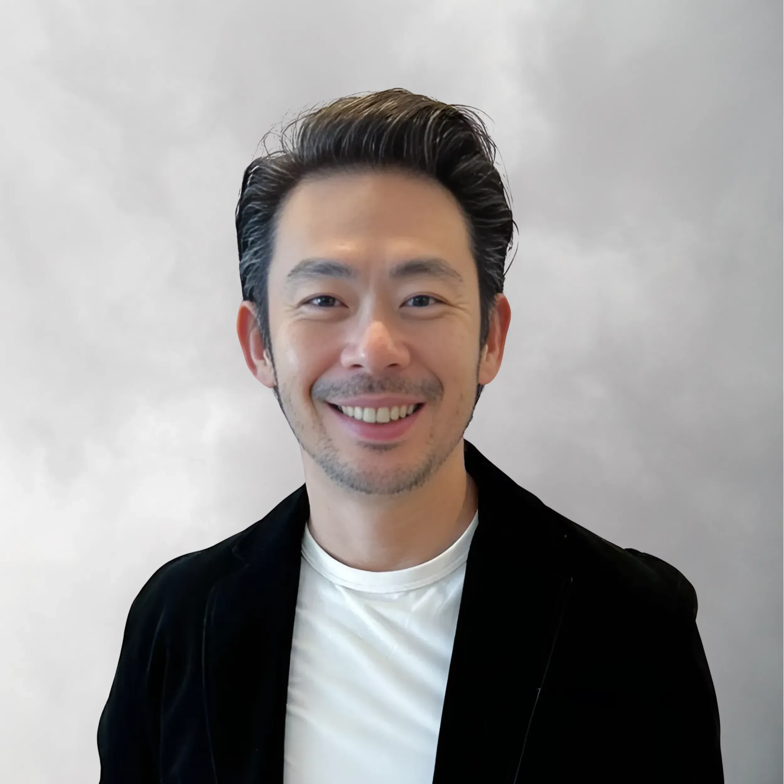 A smiling man with dark, styled hair, wearing a black jacket and white shirt, standing in front of a cloudy sky background.